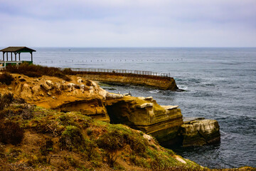 2024-12-31 THE ROCKY SHORELINE WITH THE CHILDRENS POOL IN LA JOLLA CALIFORNIA AND A CALM PACIFIC OCEAN WITH A STRING OF BROWN PELICANS FLYING ACROSS THE HORIZON AND SOFT CLOUDS