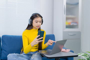 Young asian woman wearing headphones working from home sitting on a blue sofa comparing information...