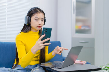 Asian freelancer wearing headphones working remotely comparing information on her laptop and...