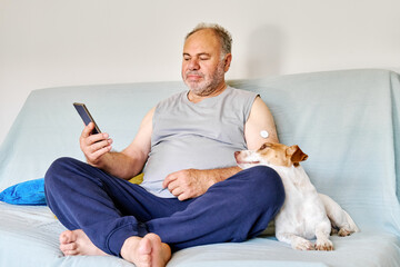 Mature man with diabetes and continuous glucose monitor on his arm using mobile app for checking his health, while sitting in sofa with Jack Russell Terrier dog at home