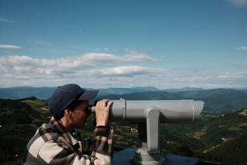 Obraz premium Woman wearing plaid shirt and cap looks through a public telescope with mountains and villages in the background, Serbia