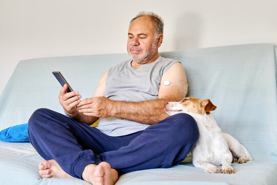 Mature man with diabetes and continuous glucose monitor on his arm using mobile app for checking his health, while sitting in sofa with Jack Russell Terrier dog at home