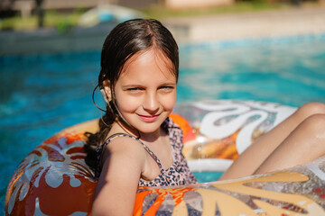 A close-up of a smiling young girl relaxing in a pool, lounging in a colorful inflatable ring. She is wearing a leopard-print swimsuit, and the sun highlights her wet hair and cheerful expression.