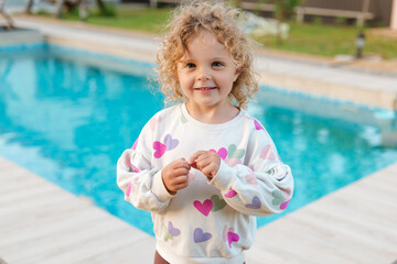 Naklejka premium Happy curly-haired girl in heart-patterned sweatshirt smiling by the pool on a sunny day, enjoying summer fun and outdoor leisure.