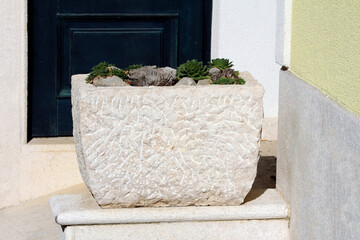 Rough-hewn stone planter holds sempervivum rosettes and rocks, placed on a step by a dark door and...