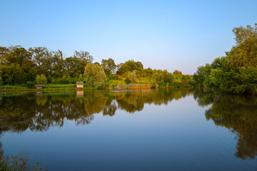 A calm lake reflects a cloudless sky and green trees, creating a harmonious summer landscape. A...