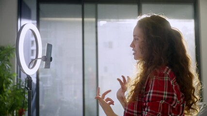 Young woman filming herself with phone and ring light during live stream session. Vlogger or content creator making reel or video for social media followers and fans in her home studio or apartment - Powered by Adobe