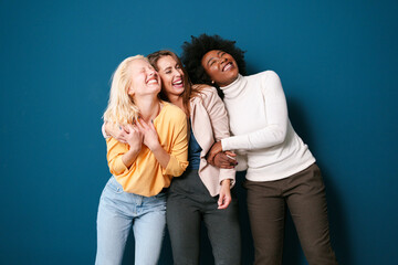 Multicultural Female Friends Sharing Authentic Laughter Moment In Casual Wear Against Deep Blue Studio Wall