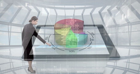 Businesswoman in dark suit manipulating floating pie chart in control room, with touchscreen tablet