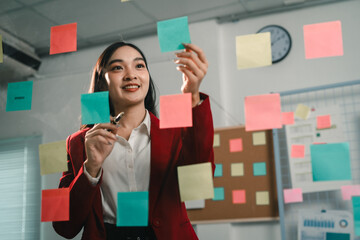Businesswoman attaching colorful sticky notes to a glass wall, organizing tasks and ideas in a...