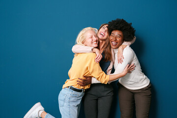 Three Multicultural Friends Sharing Laughter and Embracing in Casual Attire Against a Vibrant Blue...