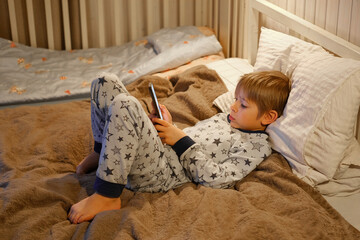Cute kid boy in pajamas lying in bed watching smartphone, smiling. Children's screen time. Child...