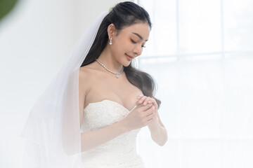 Asian Beautiful bride in elegant wedding dress smiling gently, standing gracefully in bright room,...