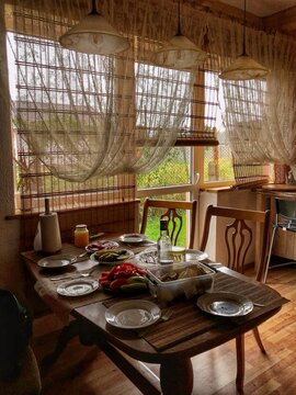 Rustic kitchen interior with a wooden table set for a family meal. Natural light coming through sheer curtains and bamboo blinds, cozy countryside atmosphere.