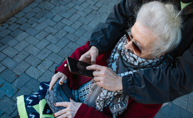 An elderly woman in a wheelchair looks at a smartphone held by a younger companion. The scene shows...