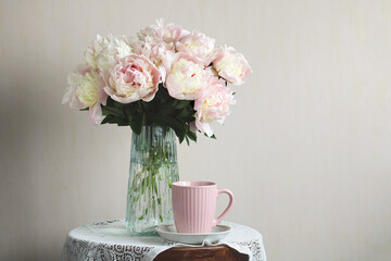 Pink and white peonies in glass vase with coffee mug on table