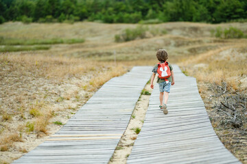 A cheerful child wearing sunglasses and a backpack running playfully on a wooden boardwalk at the Curonian Spit in Lithuania, enjoying an outdoor adventure.