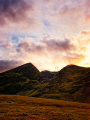 Serene sunset paints the rugged peaks of the Highlands in vibrant hues, casting a warm glow on the tranquil valley below. A solitary sheep grazes peacefully.