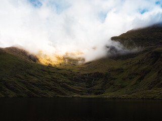 A still body of water is nestled between rolling, grass covered hills that rise into a mountain partially shrouded in white fog near Lough Callee in the Irish countryside.