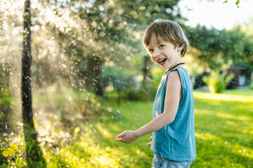 Funny little boy having fun outdoors on sunny summer day. Child running under garden sprinkler.