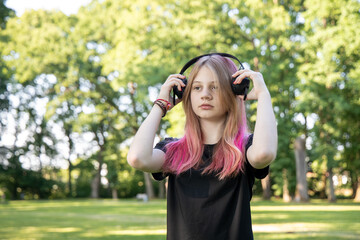 Young girl with pink hair wearing headphones stands in a lush green park, enjoying music while...