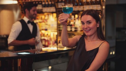 Sitting beautiful woman guest looking at camera to pose with drinking mixed vodka in blue hawaii...
