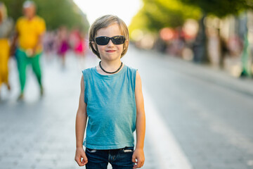 Confident young boy wearing sunglasses and a blue tank top stands in the middle of a city street on...