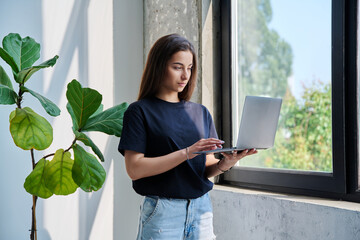 Portrait of young woman standing with laptop computer in home interior near window