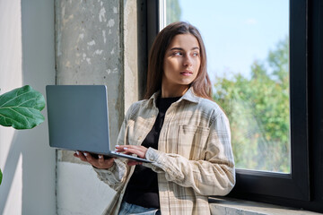 Portrait of young teenage girl using laptop standing at home, looking at window