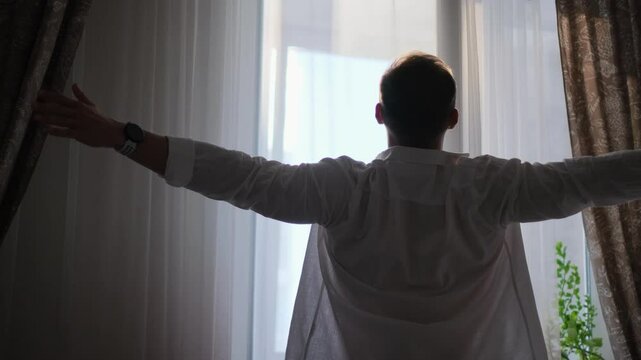 A man wakes up and stretches while standing by the window in his bedroom