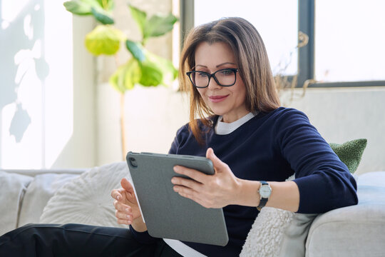 Smiling middle aged woman looking at digital tablet sitting on sofa at home