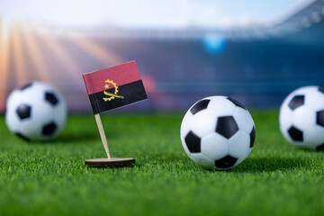 Miniature Angola flag stands prominently on vibrant green grass alongside soccer balls, symbolizing national pride and competitive spirit in a blurred stadium setting
