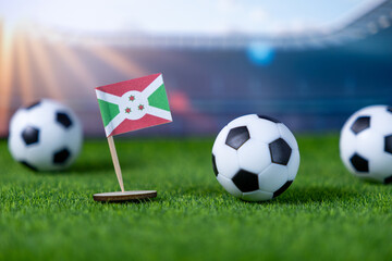 Miniature Burundi flag and soccer balls on vibrant green grass, set against a blurred stadium backdrop, representing international sports event and national pride