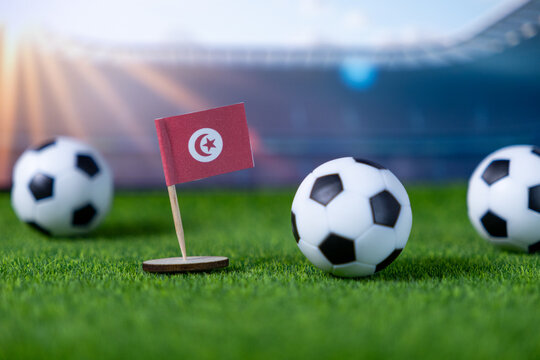 Small Tunisian flag stands proudly with soccer balls on vibrant green grass in a stadium, symbolizing national team participation in a global football competition