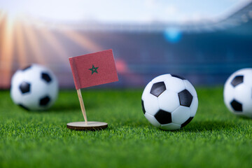 A small Moroccan flag stands with soccer balls on green grass, highlighting international football...