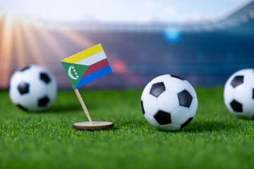 Miniature Comoros flag stands on green grass with soccer balls, symbolizing national team pride,...