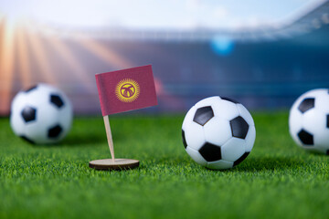 Small Kyrgyzstan flag and miniature soccer balls resting on artificial green grass with a stadium background, representing national team sports event and pride