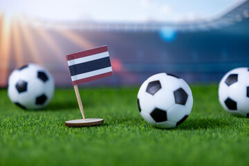 Small Thailand flag proudly stands with multiple soccer balls on lush green stadium grass, embodying national football pride and anticipation for global sports events