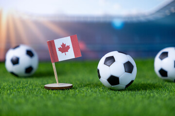 A Canadian flag and miniature soccer balls are displayed on artificial grass with a blurred stadium background, symbolizing national pride for international sports events