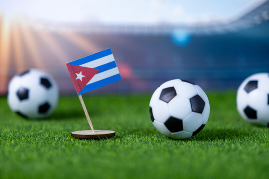 A Cuban flag stands firmly on green artificial grass with miniature soccer balls, portraying national representation and excitement for international football events in a stadium setting