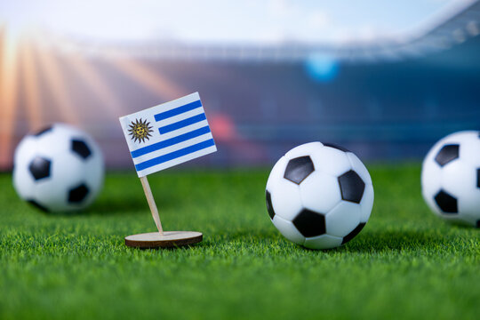 Miniature Uruguay flag displayed proudly alongside soccer balls on vibrant green grass, symbolizing international football competition and national pride in sports - Powered by Adobe
