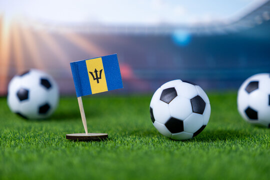 Small Barbados national flag miniature proudly stands with soccer balls on vibrant green artificial grass, symbolizing national team spirit and global football championship