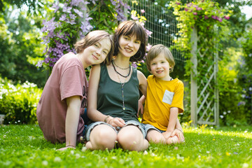 Fototapeta premium Two teenage sisters and their younger brother sit happily on the grass in a sunny garden, enjoying a warm summer day.