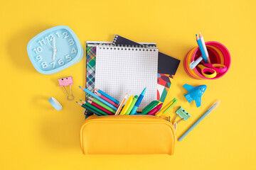 Flat lay of bright school supplies on yellow background. Stack of notebooks, alarm clock, colored pencils in yellow pencil case and various stationery. Back to school concepts.