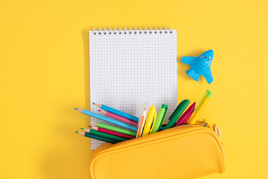Back to school concepts. Flat lay of bright school supplies on yellow background. Open notebook, full pencil case with colored pencils and airplane-shaped sharpener, top view.