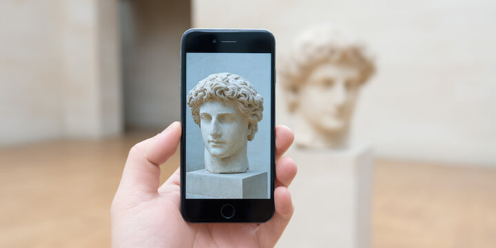 A hand holds a smartphone displaying a photo of a classical statue head, which is also visible in the background. Concept: blending technology and nft art
