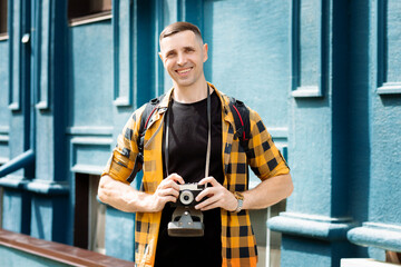 A young man with a short haircut, in a checkered shirt, holds a camera, smiles, stands against the...