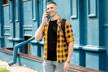 Fototapeta premium Young man in yellow checkered shirt talking on phone, smiling, wearing backpack, watch and jeans. Background is blue building.