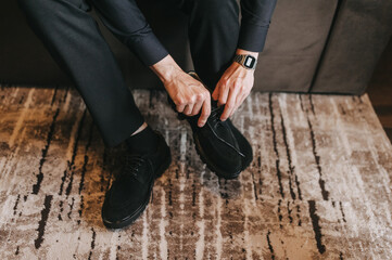 Young male businessman putting on black leather shoes on his foot in the morning, tying laces. Wedding photography.
