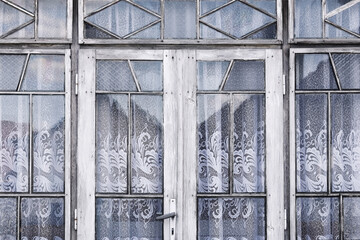 Old wooden door with glass windows. Decorative glass with lace curtains pattern. Vintage entrance white peeling paint. Retro architecture element. Rustic style detail. Traditional house window view.